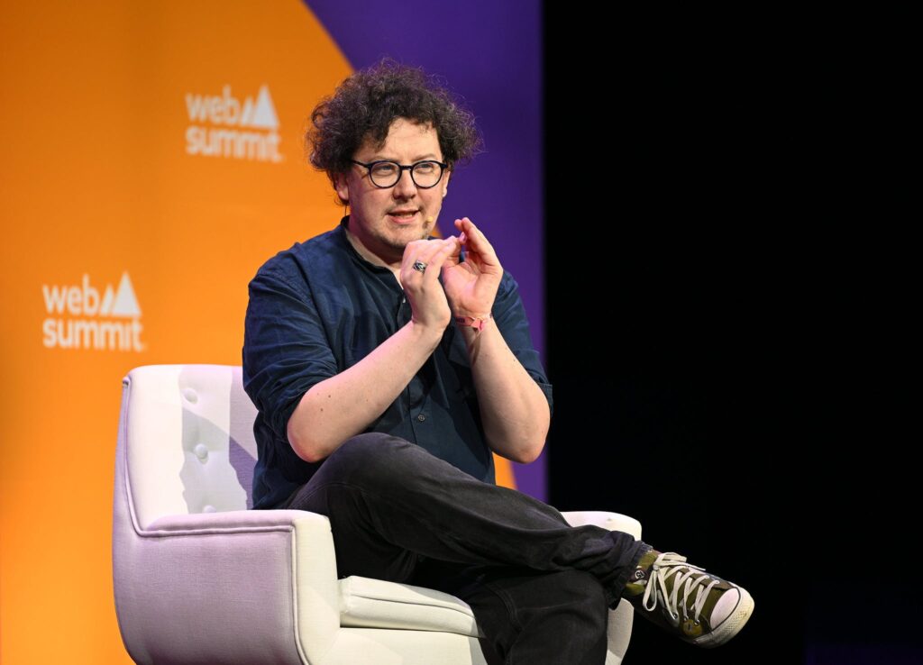 A person sits in an armchair, speaking and clasping their hands together. The background is a large circle on a solid background.