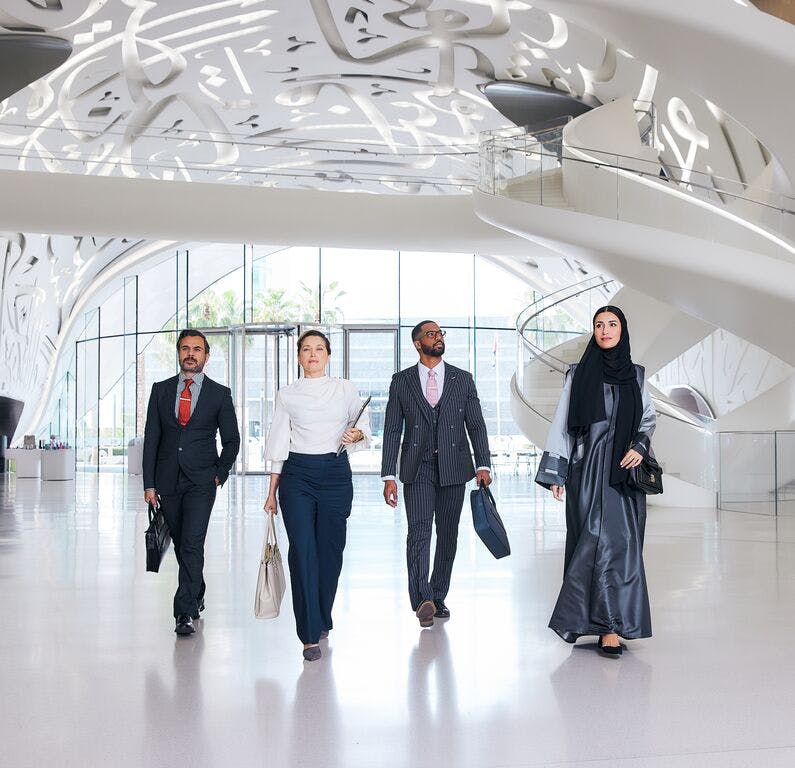 Four people work purposefully towards the lens. Each is carrying a laptop bag or briefcase. One of the people is carrying a closed laptop in their left hand, balancing it on their hip. They are in a spacious glass atrium, which appears to be the lobby of an office building.