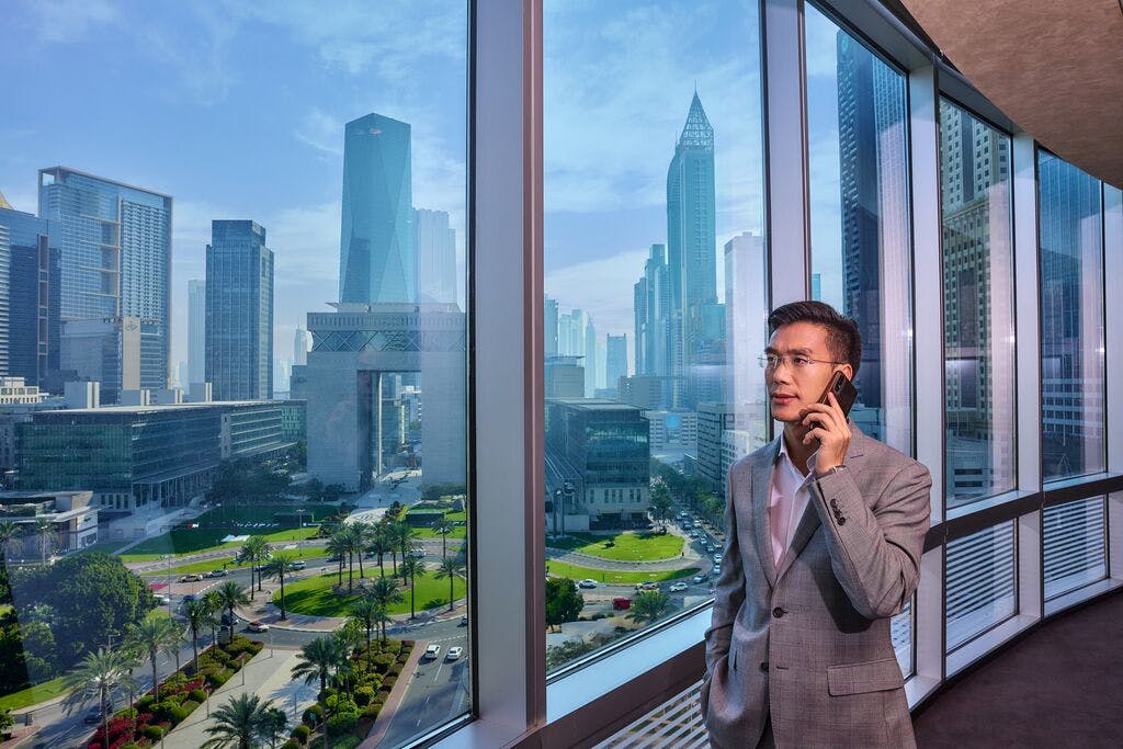 A person holds a smartphone to their ear. They stand next to a wall of windows, which overlook a modern cityscape boasting dozens of high-rise buildings. In the foreground of the cityscape is a smaller multi-storey building in the shape of an arch. This arch-style building is surrounded by even lower multi-storey, glass-fronted buildings. There are several palm trees in the centre of a roundabout in front of the arch-style building. This is Dubai.