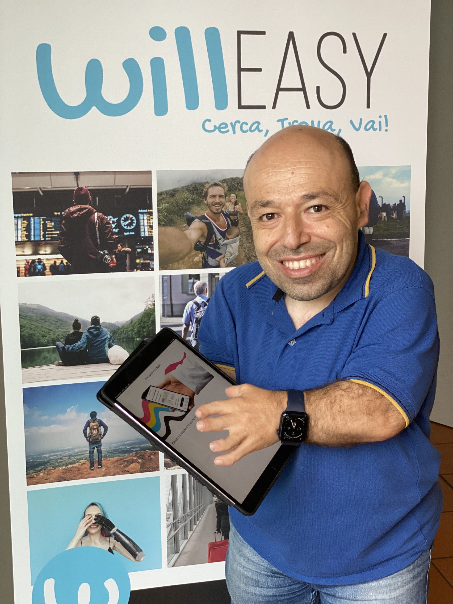 A person stands in front of a photo wall that features photos of people travelling and of people with disabilities. Text on the wall reads 'Willeasy. Cerca, Trova, Vai!' The person is holding a tablet computer.